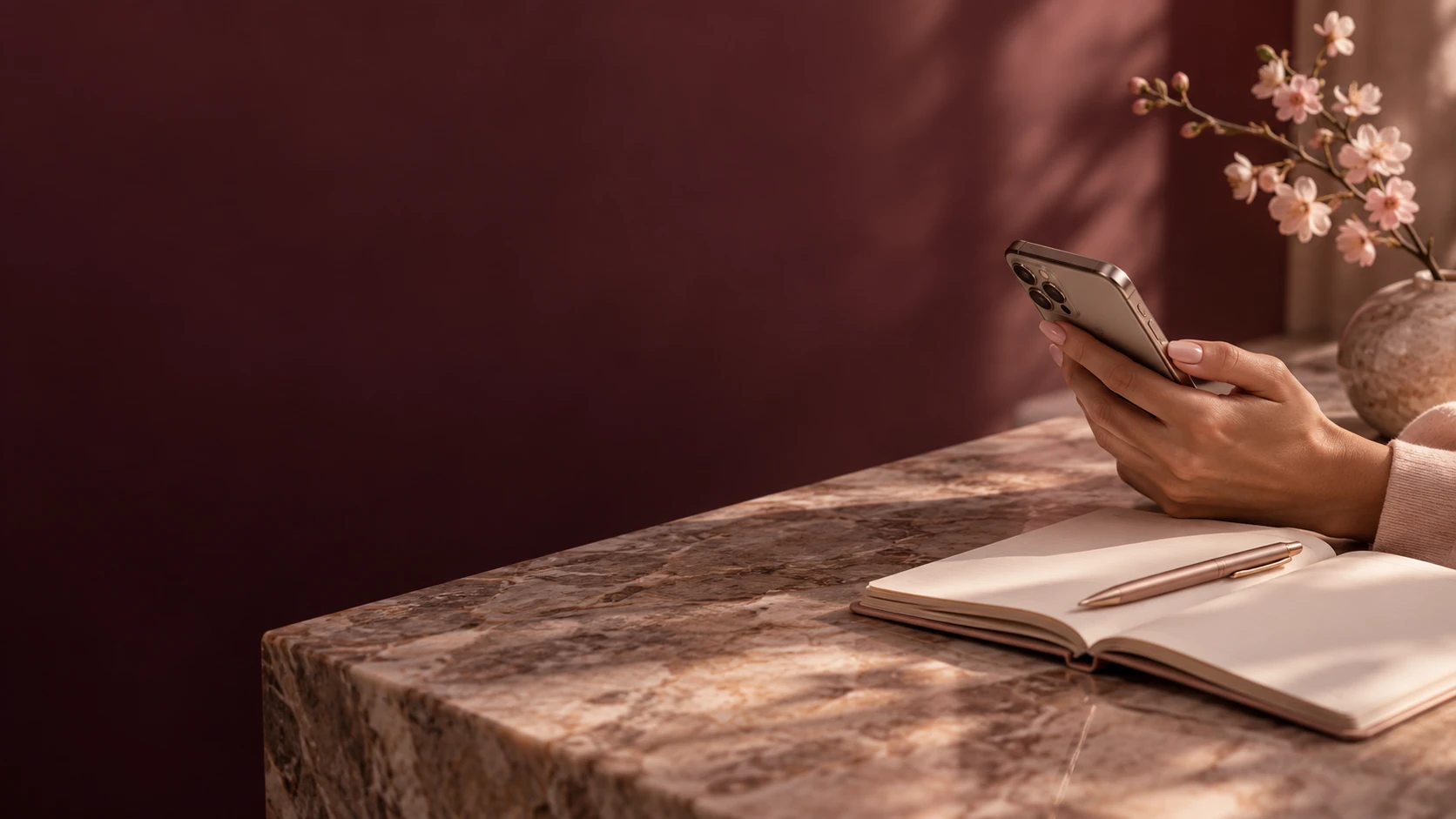 Hand holding a smartphone above an open notebook on a stone desk with pink blossoms