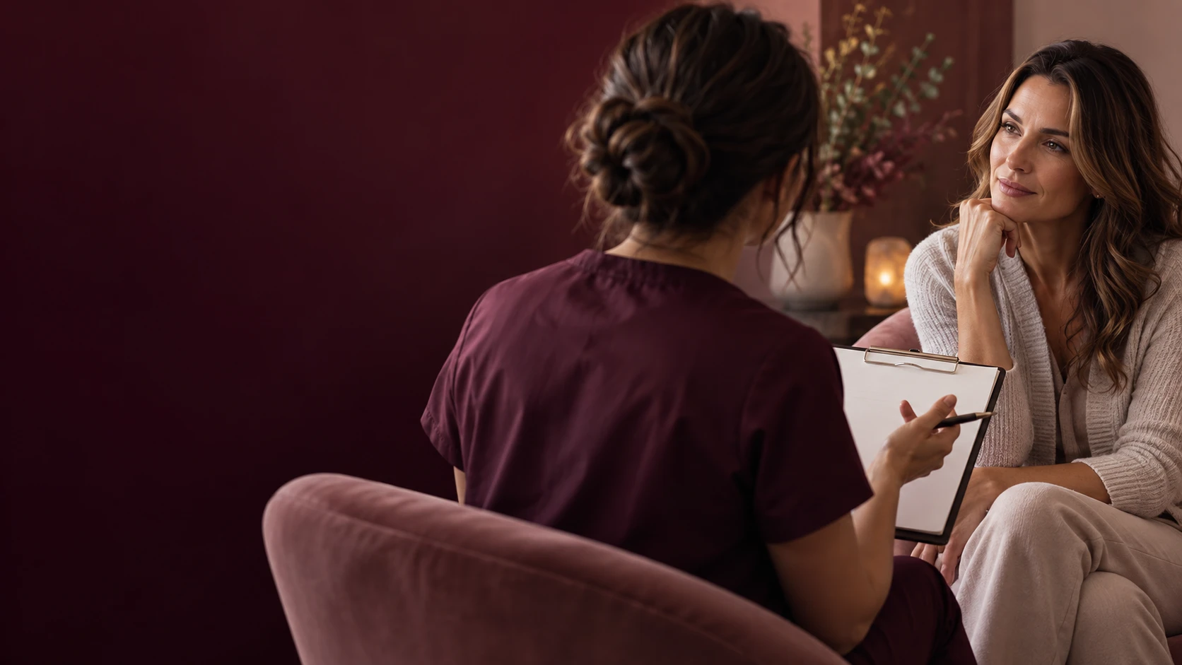 Provider taking notes on a clipboard while talking with a woman seated in an armchair