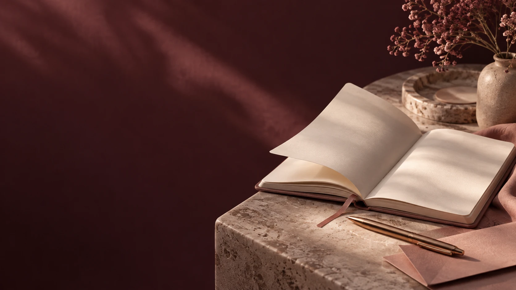 Open notebook and rose-gold pen on a stone table beside a vase of dried pink flowers