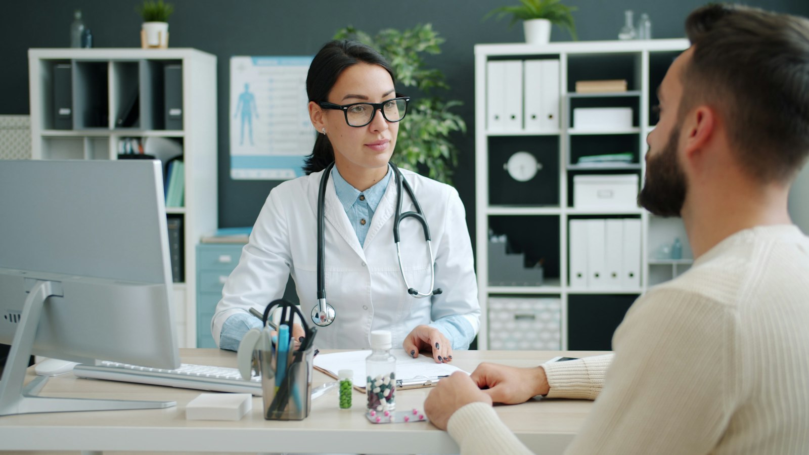 Consultation room — provider in a white coat and glasses talking with a patient across a desk